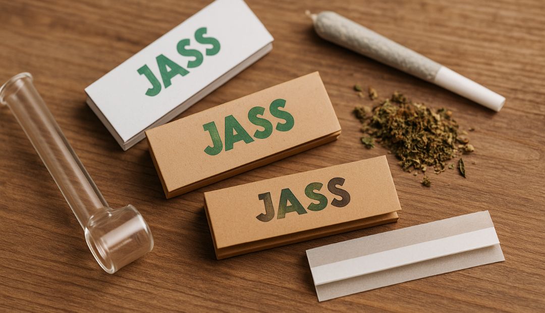 Packs de feuilles à rouler Jass marron et blanches posés sur une table en bois, avec un joint roulé, du CBD effrité et un accessoire en verre